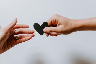 Image of two hands against a white background. One of the hands is passing a black paper heart to the other one